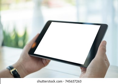 Mockup Image Of Hands Holding Black Tablet Pc With Blank White Screen On Wooden Table In Cafe