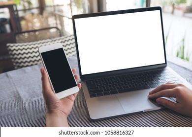 Mockup Image Of Hands Holding Blank Mobile Phone While Using Laptop With Blank White Desktop Screen On Table 