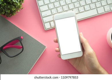 Mockup Image Of Hands Holding White Mobile Phone With Blank White Screen With Modern Pink Office Desk With Laptop And Other Supplies For Input The Text On Copy Space Top View, Flat Lay.