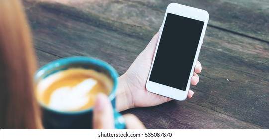 Mockup Image Of Hands Holding White Mobile Phone With Blank Black Screen And Hot Latte Art Coffee On Vintage Wood Table In Outdoor Park