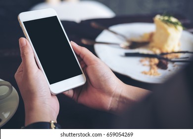 Mockup Image Of Hands Holding White Mobile Phone With Blank Black Screen With A Plate Of Cake On Wooden Table In Restaurant