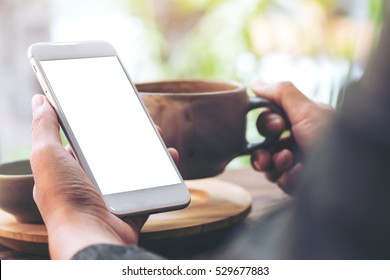 Mockup Image Of Left Hand Holding White Mobile Phone With Blank White Screen And Right Hand Holding Hot Latte Art Coffee Cup While Looking And Using It At Vintage Wooden Table In Cafe