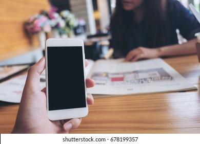 Mockup Image Of A Man's Hand Holding White Mobile Phone With Blank Black Screen In Modern Cafe And Blur Woman Reading Newspaper In Background