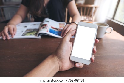 Mockup Image Of A Man's Hand Holding And Using White Smart Phone With Blank Screen On Wooden Table With Woman Reading Magazine In Background