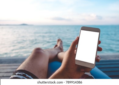 Mockup image of a man's hand holding white mobile phone with blank desktop screen sitting by the sea and blue sky background - Powered by Shutterstock