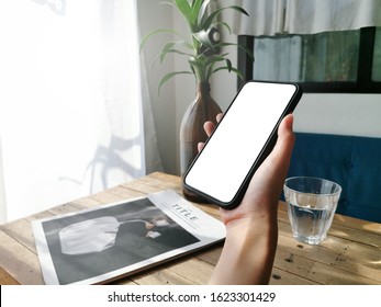 Mockup image on the hand that is holding a mobile phone with white screen paths and mockup magazine book at a local wooden table in a cafe. 