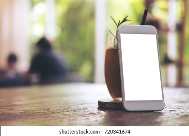 Mockup Image Of White Mobile Phone With Blank Screen With A Glass Of Iced Coffee On Wooden Table In Modern Cafe With Blur Green Nature Background