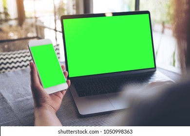 Mockup Image Of A Woman Holding Blank Mobile Phone While Using Laptop With Blank Green Screen On Table 