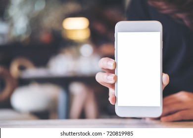 Mockup image of a woman holding and showing white mobile phone with blank screen on the table in modern cafe