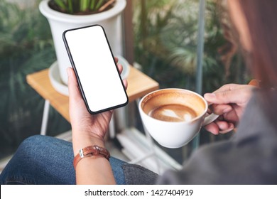 Mockup image of a woman holding and using black mobile phone with blank desktop screen while drinking coffee in cafe