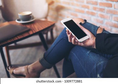 Mockup Image Of A Woman Sitting Cross Legged , Holding White Mobile Phone With Blank Black Desktop Screen On Thigh In Cafe