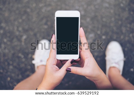 Mockup image of a woman sitting on the street and holding white mobile phone with blank white screen   