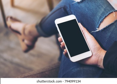 Mockup Image Of A Woman's Hand Holding White Mobile Phone With Blank Black Desktop Screen On Thigh In Cafe