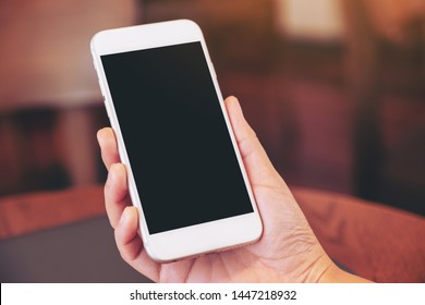 Mockup Image Of Woman's Hands Holding Black Mobile Phone With Blank White Screen With Laptop Computer On Wooden Table