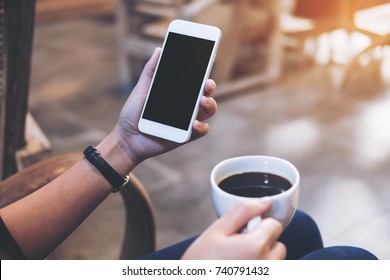 Mockup image of woman's hands holding white mobile phone with blank black screen while drinking coffee in modern loft cafe