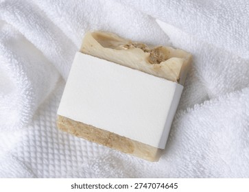 Mockup of light beige solid soap bar with blank paper label on white bath terry towels. Rectangular shape. Unbranded packaging on neutral background with copy space, top view. Personal care context