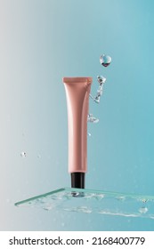 Mockup Of A Pink Tube With Cream On A Glass Shelf And Splashes Of Water On A Blue Background Close-up. Place For A Logo.