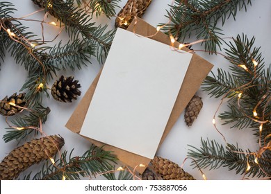 Mockup With Postcard And Branches Of A Christmas Tree And Cones On A White Background With A Garland. Winter Festive Mood.