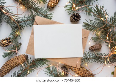 Mockup With Postcard And Branches Of A Christmas Tree And Cones On A White Background With A Garland. Winter Festive Mood.