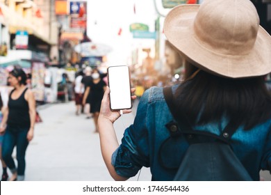 Mockup Of Smart Phone Blank White Screen. Holding Hand By Back Shoulder View Of Traveller Asian Woman With Hat And Backpack. Famous Destination Background Travel In Bangkok, Thailand.