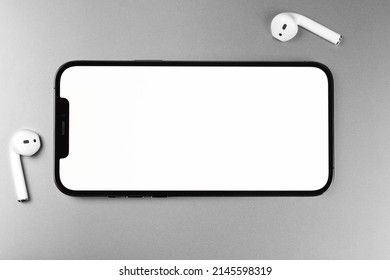 Mockup Smartphone IPhone Horizontal With White Screen, Empty Display With AirPods Headphones. Apple Is A Multinational Technology Company. Batumi, Georgia - March 19, 2022