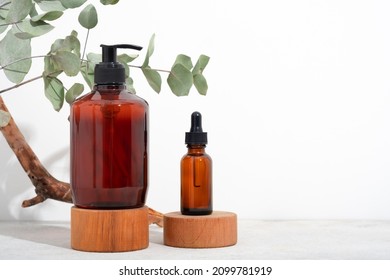 Mockup Spa Body Cosmetics In Brown Glass Bottles Serum With A Pipette, Oil, Gel, Shower Soap, Shampoo With A Pump On A Wooden Pedestal And A White Background With Eucalyptus Branches. Copy Space