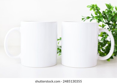 Mockup of two white coffee mugs on table with greenery. Blank 11 oz ceramic cups mock up.