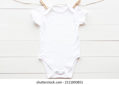 Mockup Of White Baby Bodysuit On Wood Background With Clothespins And Rope, Flat Lay. Blank Gender Neutral Baby Clothes Template In Farmhouse Style.