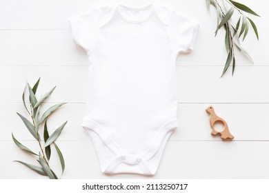 Mockup of white baby bodysuit on wood background with greenery and toy. Blank baby clothes template, flat lay.
