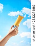 A mockup yellow cream tube is being held by a female model against a clear blue sky with white clouds drifting by. Minimalist image used for advertisements of skincare products.