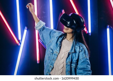 Mod curly dark haired girl dressed in blue denim jacket uses the virtual reality glasses on her head in the dark studio with neon light - Shutterstock ID 1620999430