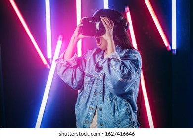 Mod curly dark haired girl dressed in blue denim jacket uses the virtual reality glasses on her head in the dark studio with neon light  - Shutterstock ID 1627416646