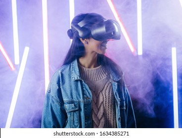 Mod curly dark haired girl dressed in blue denim jacket uses the virtual reality glasses on her head in the dark studio with neon light and smoke fog - Shutterstock ID 1634385271
