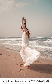 Model in boho style in a white long dress and silver jewelry on the beach. Her hair is braided, and there are many bracelets on her arms. - Shutterstock ID 2256469579