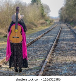 Model with her back to the camera standing on the train tracks, a guitar and pink shawl on her back, transparent black dress, pink shawl, brown hat and high heels, autumn day with blurred background