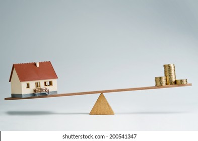 Model House And Money Coins Balancing On A Seesaw