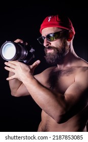 Model With A Naked Torso With A Camera In Her Hands. The Photographer Has A Red Cap And Green Glasses On His Head. Guy On A Black Background