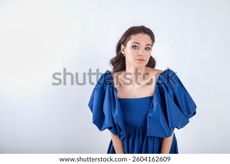 A model poses indoors in a studio setting, wearing a striking blue dress with puffy sleeves. The plain backdrop emphasizes the outfit’s vibrant color and design, creating a stylish ambiance.