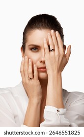 A model poses in a studio with a neutral background, partially obscuring her face with one hand while showcasing rings on her fingers. Her expression is serene and captivating.