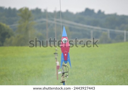 A model rocket imitating a steampunk rocketship with a martian inside. It is mounted on a launch pad and is red with blue fins with white stars. The background is a green field and green trees.