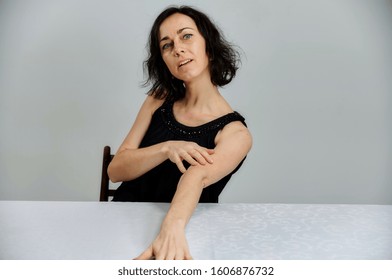 Model Sits at a table right in front of the camera with vivid emotions. Portrait of a cute smiling talking brunette woman in a black dress on a white background.