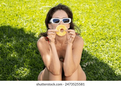 Model Sitting On The Grass, Facing Forward, Holding A Pineapple Slice In Front Of Her Mouth As If It Were Her Smile. She Wears A Red And Black Bikini, White Sunglasses, And Red Lipstick, With Freckles