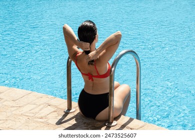 Model sitting on the pool stairs, drying her hair from the back, holding it with both hands. She exudes a sense of relaxation and tranquility in the poolside setting - Powered by Shutterstock