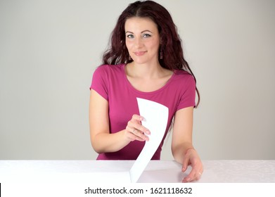 The Model Smiles And Makes A Financial Report. Portrait Of A Pretty Slender Brunette Woman In A Pink Dress And With Brown Hair Sitting At A Table On A White Background In The Studio.