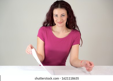 The Model Smiles And Makes A Financial Report. Portrait Of A Pretty Slender Brunette Woman In A Pink Dress And With Brown Hair Sitting At A Table On A White Background In The Studio.