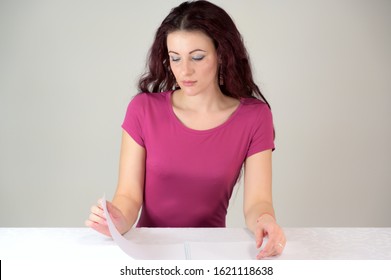 The Model Smiles And Makes A Financial Report. Portrait Of A Pretty Slender Brunette Woman In A Pink Dress And With Brown Hair Sitting At A Table On A White Background In The Studio.