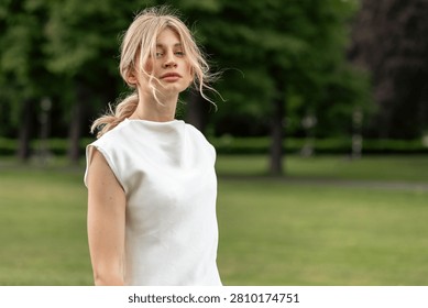 Model stands in park with trees in background during bright day while wearing simple white outfit and looking toward camera