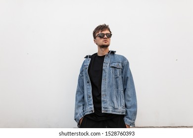 Model of a stylish young man with a stylish hairstyle with a beard in fashionable sunglasses in trendy clothing near a vintage white wall in the city. Attractive hipster guy outdoors . Youth fashion.