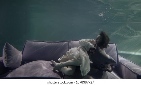 A Model Woman In A Dressing Gown Is Lying Posing On The Back Of A Gray Sofa Holding A Pillow In Her Hands, The Sofa Is Under Water, It Reflects The Light From The Waves.