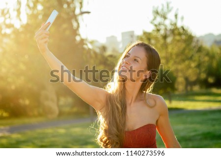 model woman long-haired in red dress does selfie on the phone rays of sun park background